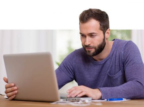 A bearded man sitting at a table, working on a laptop.