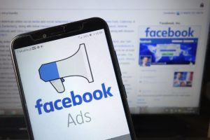 A smartphone displaying the "Facebook Ads" logo in the foreground, with a computer screen showing the Facebook website in the background.