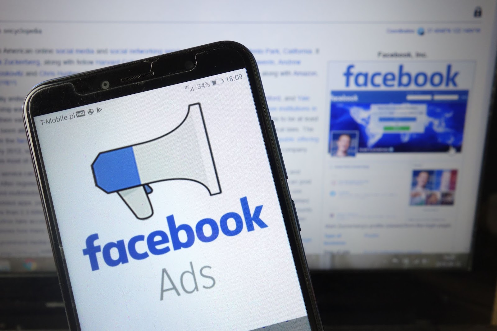 A smartphone displaying the "Facebook Ads" logo in the foreground, with a computer screen showing the Facebook website in the background.