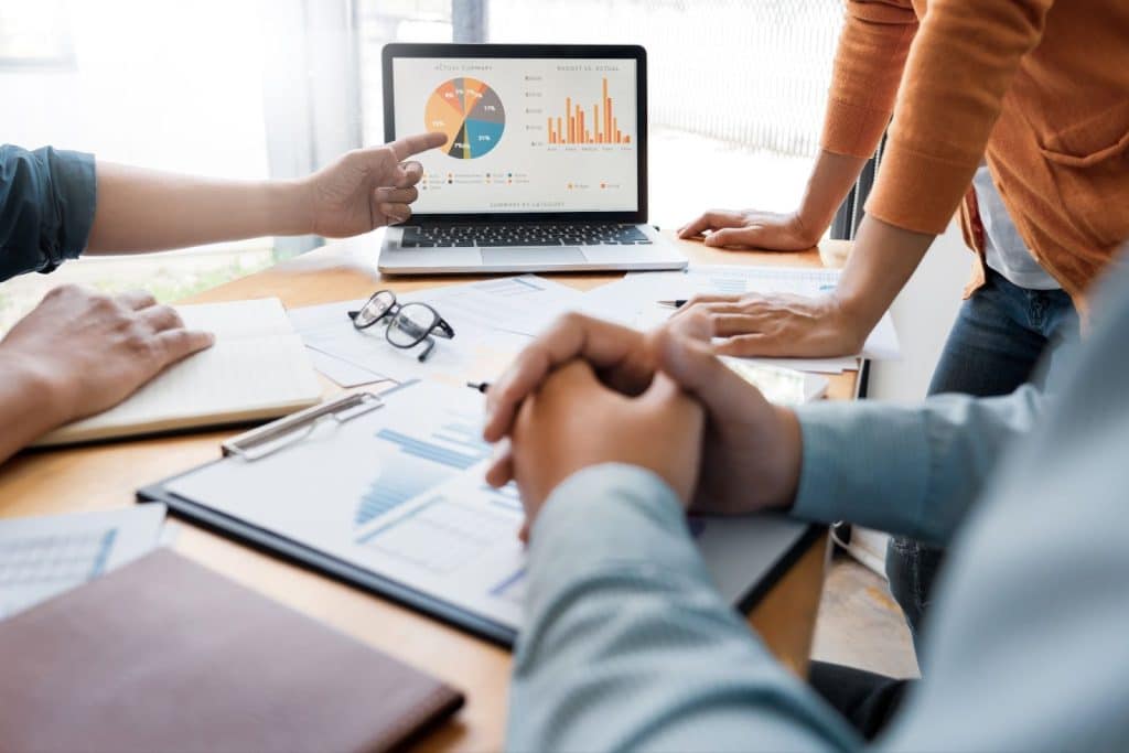 Team of business individuals engaged with a laptop, reviewing graphs that illustrate marketing strategies and sales objectives.