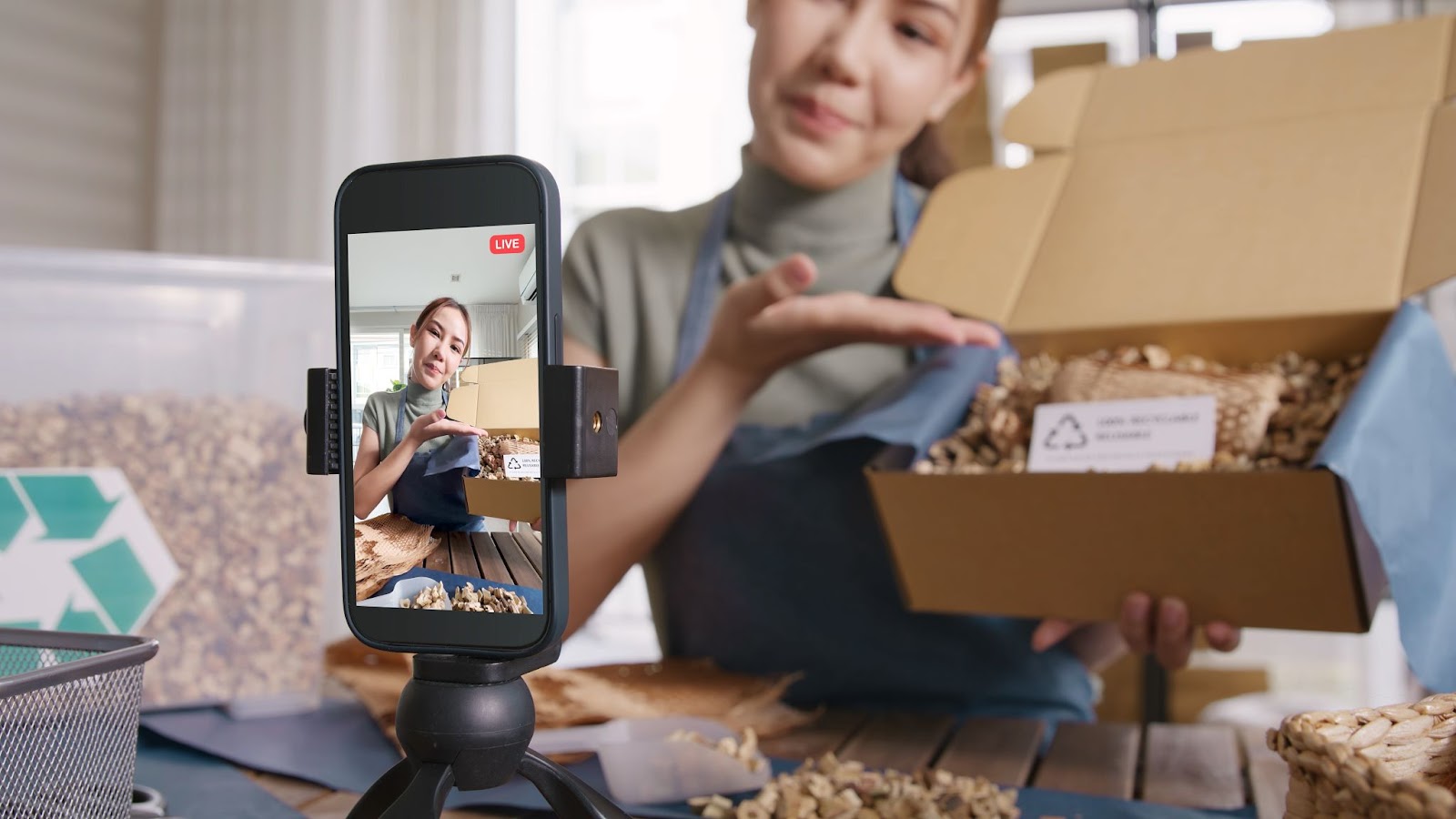 A female influencer livestreaming an unboxing video, presenting a sustainable packaging product. The phone on a tripod captures her demonstration, showing an eco-friendly package with recyclable materials.