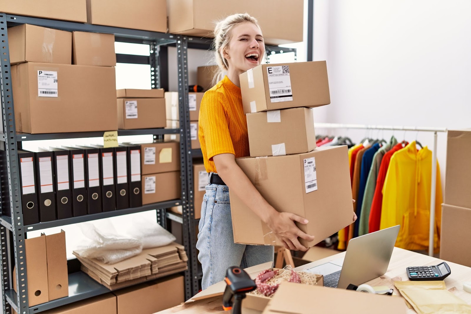 Person in a warehouse holding a stack of labeled boxes, surrounded by shelves and clothing racks.