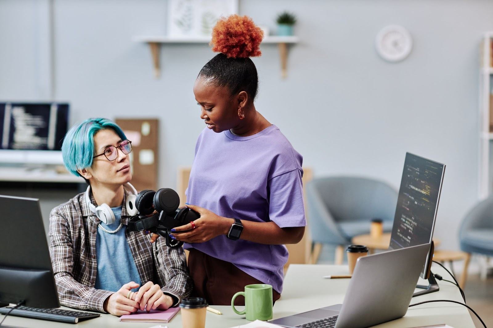 Two people in an office discuss a camera setup at a desk with laptops and coffee mugs.