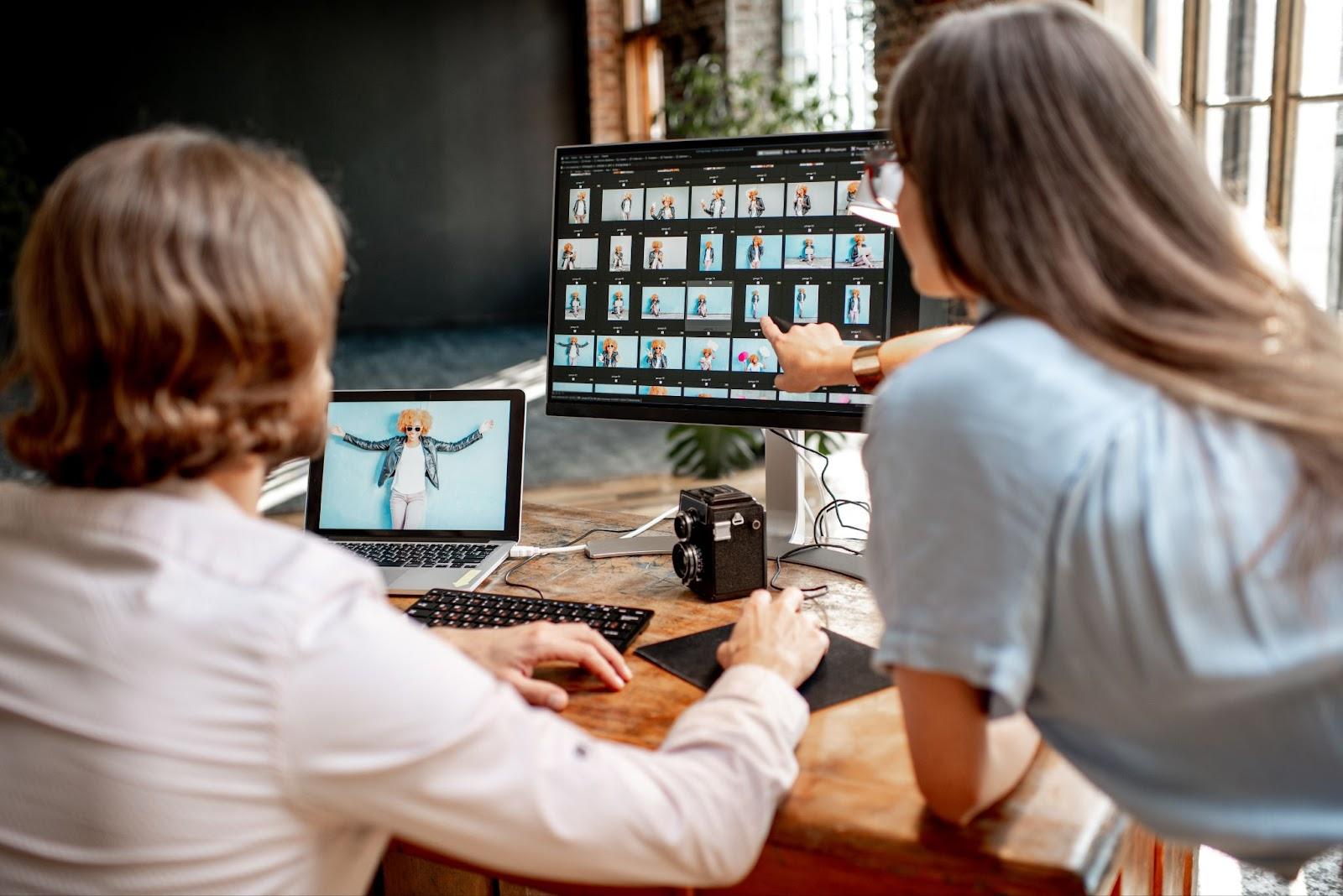 Two people review photos on a computer and laptop at a wooden desk with a camera.