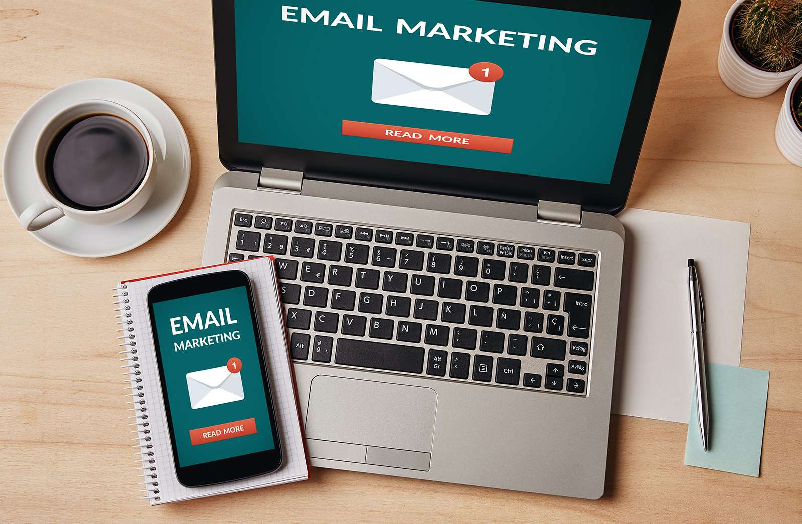 Laptop and smartphone displaying email marketing screens, with coffee cup, notebook, and pen on a wooden desk.