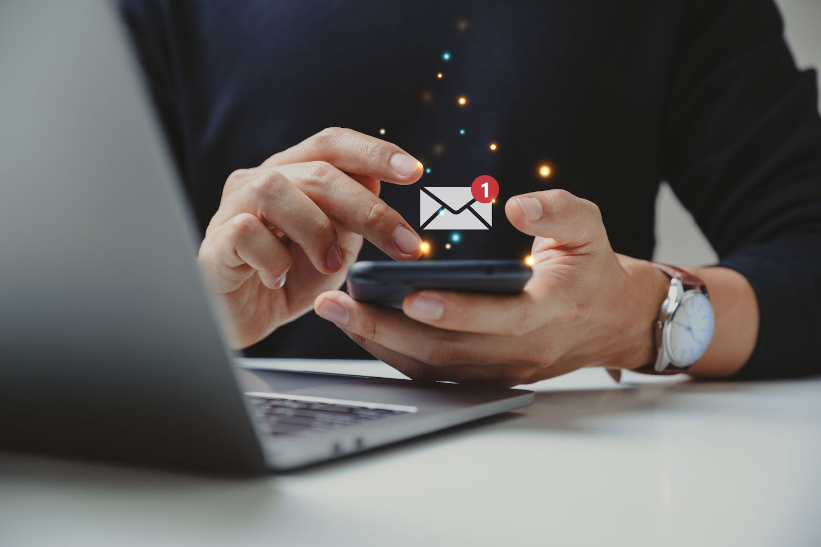 A person using a smartphone with a new email notification icon, while working on a laptop, with a watch on their wrist.