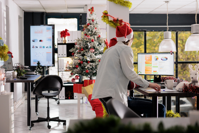 Person in a Santa hat working at a desk with a computer displaying charts, surrounded by Christmas decorations and a tree.
