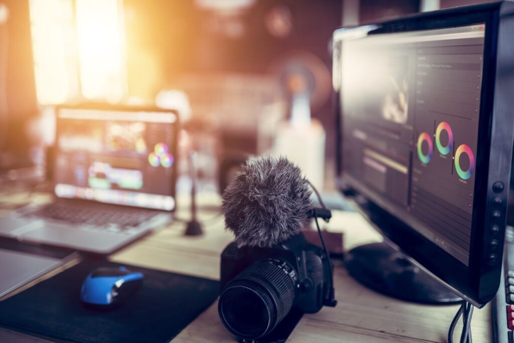 A workspace with a camera, microphone, laptop, and monitor displaying color wheels, bathed in warm sunlight.