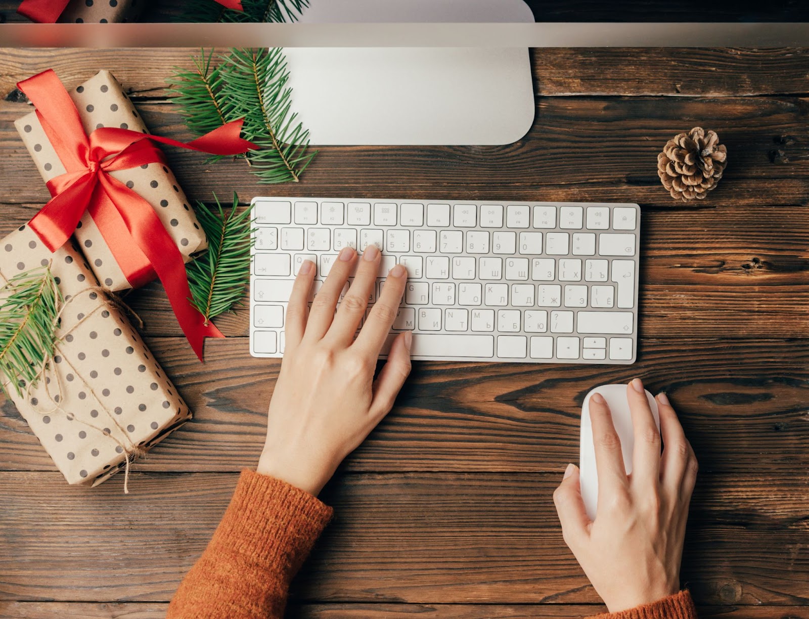 Person typing on keyboard with Christmas gifts and pine branches on wooden desk.