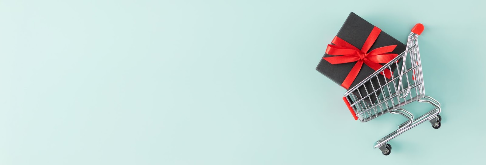 Shopping cart with black gift box tied in red ribbon on mint background