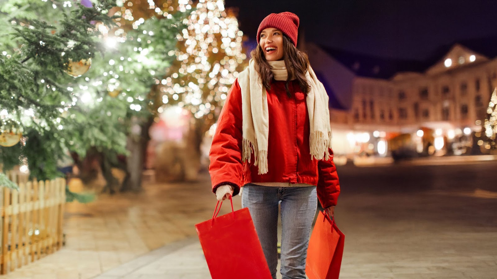 Smiling woman in red coat and beanie carries red shopping bags by glowing Christmas trees at night.