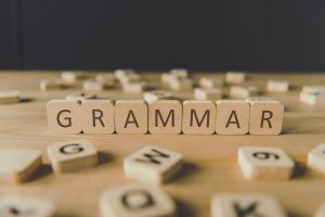 Wooden letter blocks spelling GRAMMAR on a table, with scattered alphabet tiles in the background. Focus on the word in the foreground.