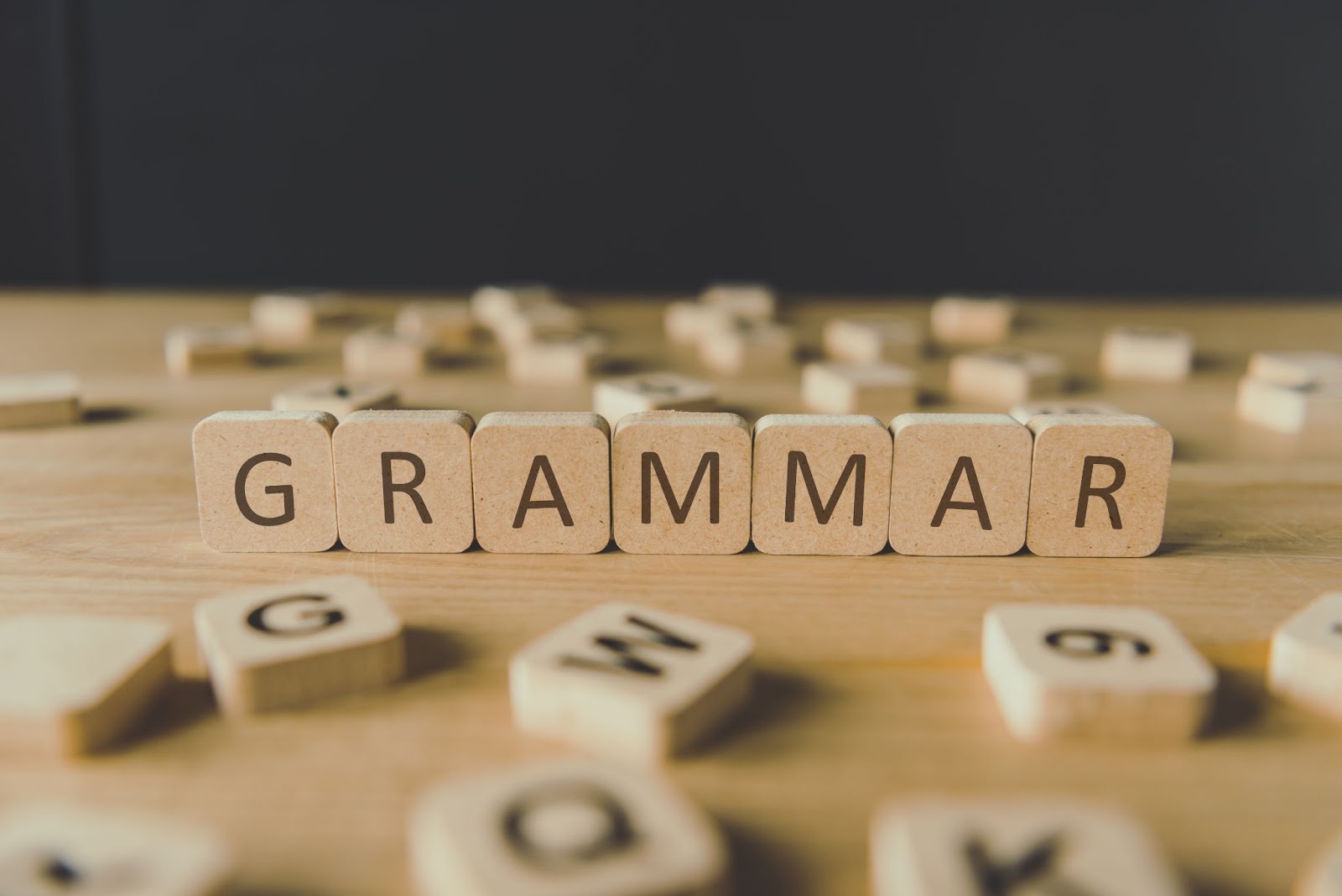 Wooden letter blocks spelling GRAMMAR on a table, with scattered alphabet tiles in the background. Focus on the word in the foreground.