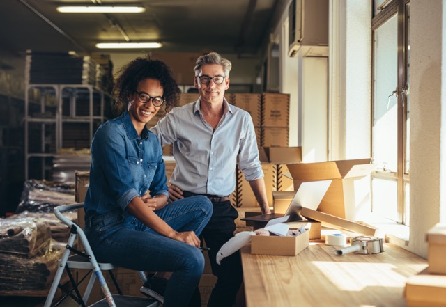 A man and a woman smiling in a warehouse setting, surrounded by cardboard boxes and shipping supplies.