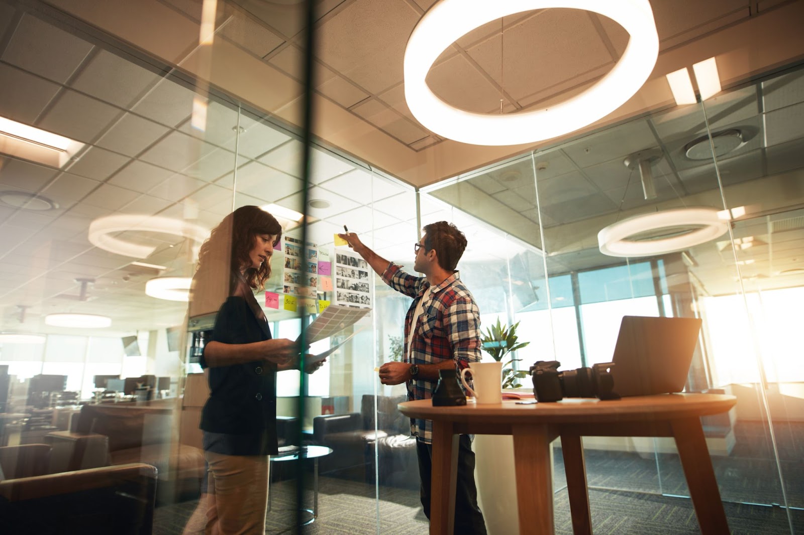 In an office setting, two individuals converse under a circular light fixture, highlighting a professional atmosphere.
